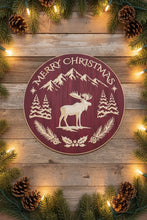 A round wooden Christmas sign with a dark red background and light natural wood cutout details, featuring a moose, mountains, pine trees, holly, and the words 'MERRY CHRISTMAS'. The sign is photographed on a rustic wood bwnch with Christmas lights