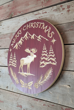 A round wooden Christmas plaque with a dark reddish-brown finish and light-colored laser-cut details of a moose, pine trees, mountains, and the text 'MERRY CHRISTMAS'.